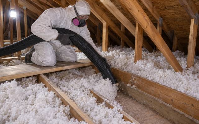 Worker in a white suit and respirator vacuuming loose white fiberglass out of an attic with a black hose