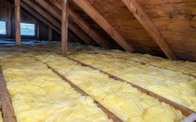 Yellow fiberglass batts between joists in an older attic with board sheathing and a louvred gable vent